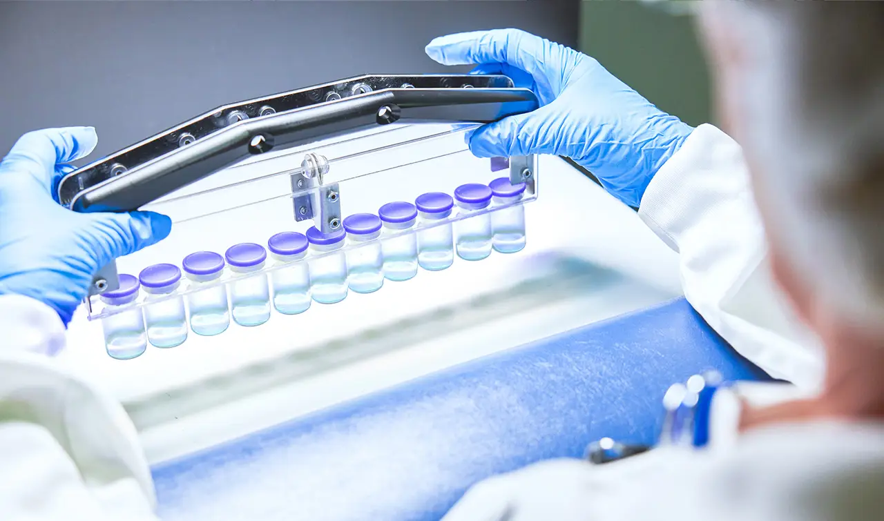 Specialist in blue gloves performing a visual quality inspection of a rack of clinical drug product vials.